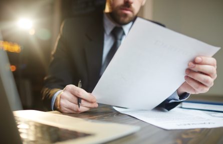 Portrait of modern bearded businessman holding papers in hands, reading and analyzing contract documentation at desk with laptop